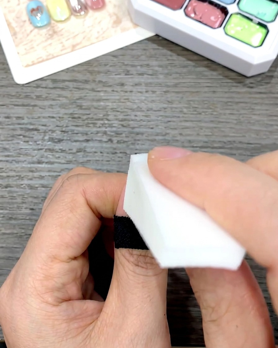 Una mano usando una esponja cosmética blanca en forma de cuña para dar toques suaves y difuminar el esmalte de gel amarillo hacia abajo en una uña.