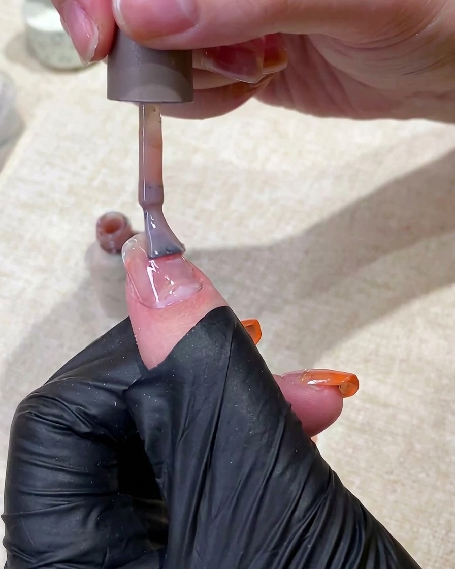 Una mano con guante negro usando un pincel para aplicar una capa base de gel nude translúcido en una uña.