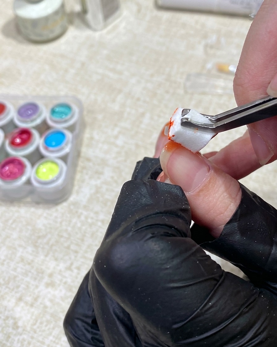 Una mano enguantada usando pinzas para aplicar gel naranja desde una pequeña esponja en la punta de una uña.