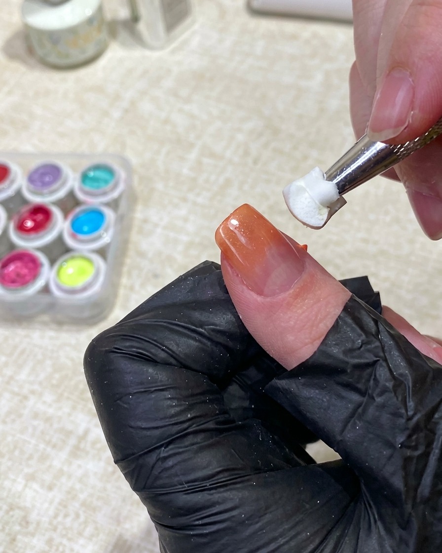 Una herramienta sosteniendo una pieza de esponja blanca limpia, dando toques suaves en el medio de una uña para difuminar el gel naranja en la base nude.