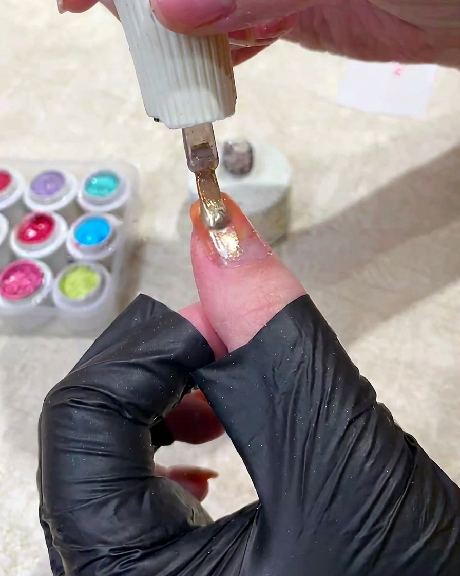 Un pincel de uñas aplicando un esmalte de gel cat eye dorado brillante sobre la uña con degradado naranja.