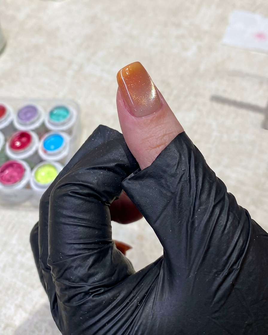 Una mano con guante negro usa pinzas para sostener un aplicador de esponja pequeño, aplicando suavemente gel naranja brillante en la punta de una uña manicurada.