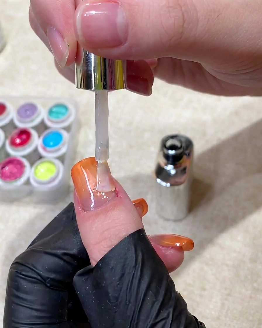 Un pincel aplicando un top coat claro y brillante sobre una uña terminada con degradado cat eye naranja.