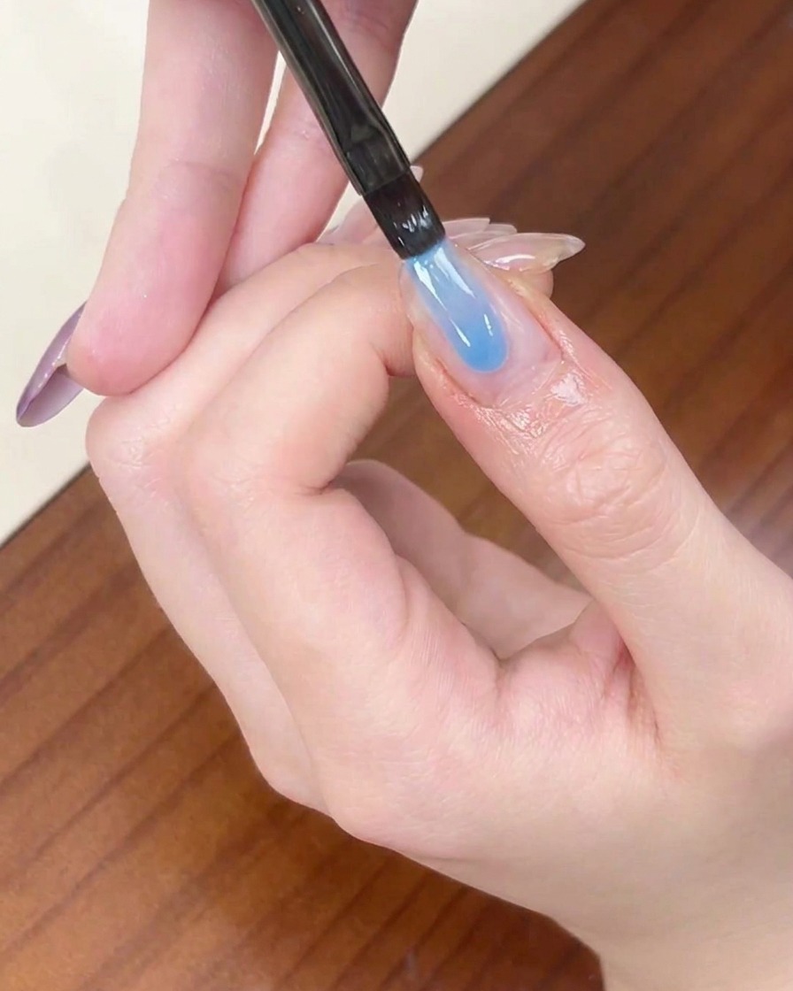 Un pincel delineador aplicando una franja de gel azul transparente en la punta de una uña almendrada.