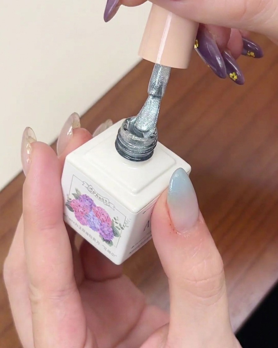 Un pincel de esmalte en gel aplicando una capa de esmalte ojo de gato plateado brillante sobre una uña con degradado azul.