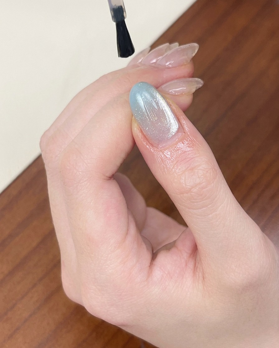 Un pincel aplicando un top coat transparente y brillante sobre una uña con gel ojo de gato azul claro.
