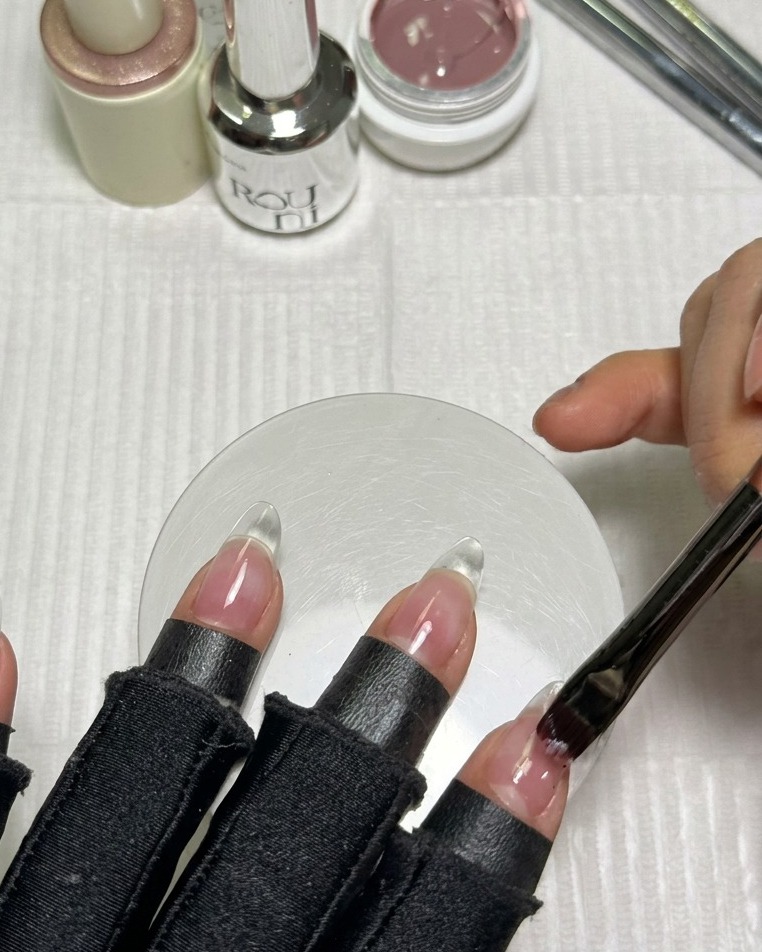 Un pincel aplicando una capa uniforme de esmalte en gel nude rosado sobre una extensión de uña transparente con forma almendrada.