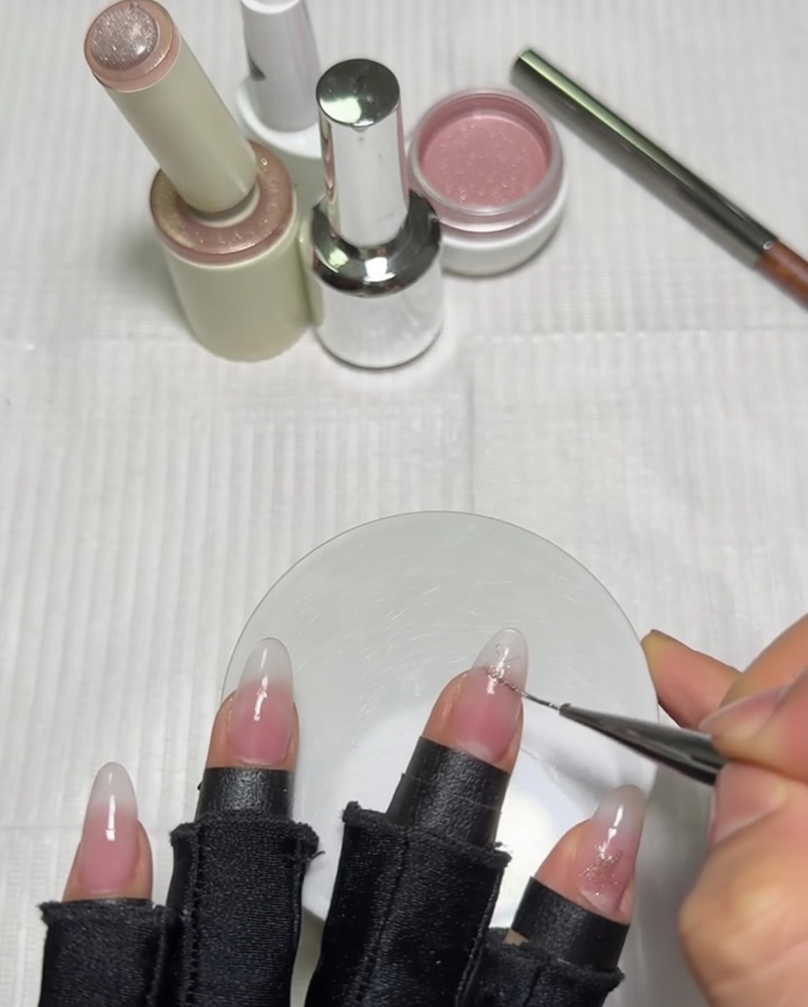 Primer plano de una mano usando un pincel delineador largo para detallar un diseño de estrella rosa en la uña del pulgar que ya tiene una base degradada lechosa.
