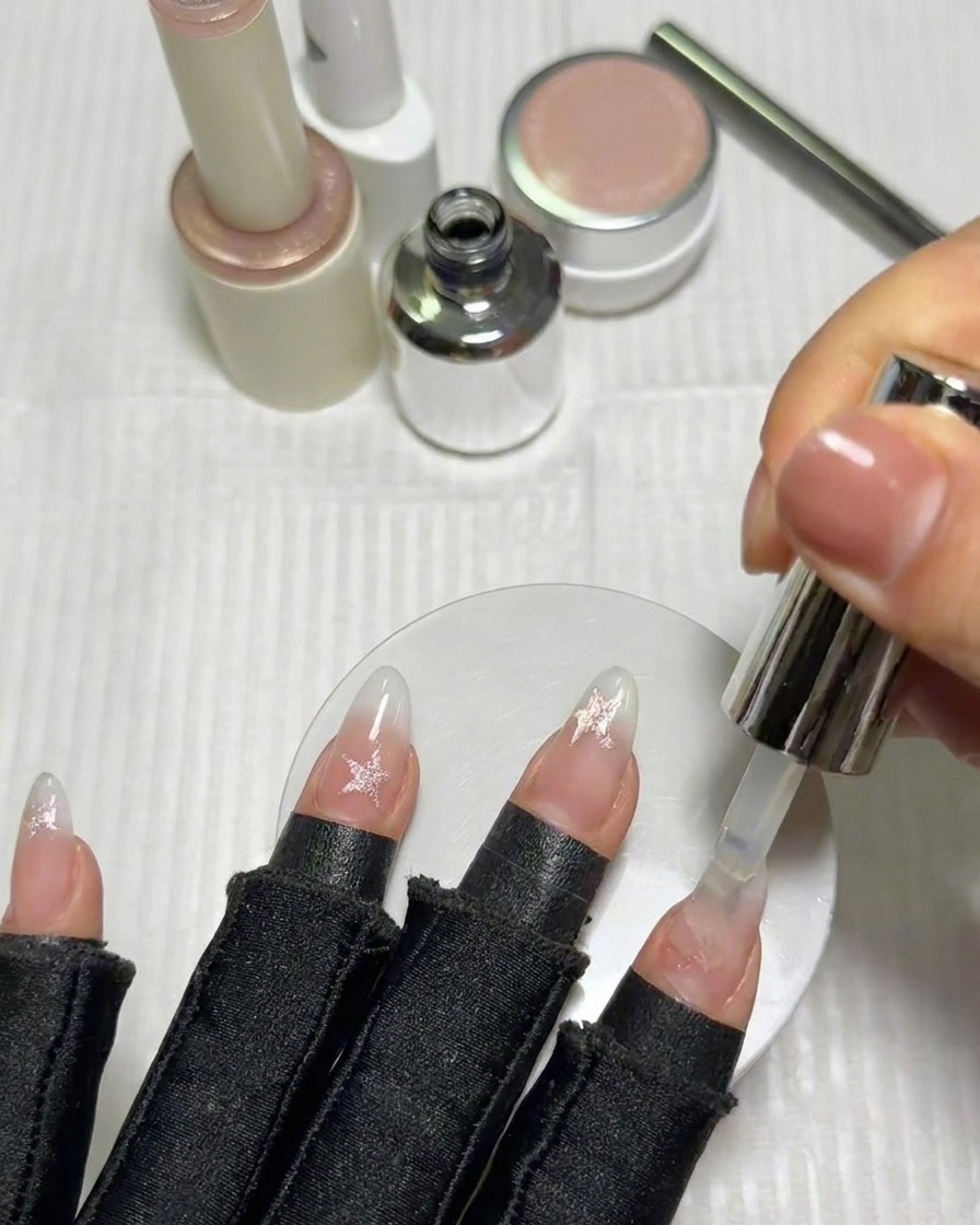 Un pincel aplicando una capa final de brillo transparente sobre las uñas terminadas y decoradas con estrellas.