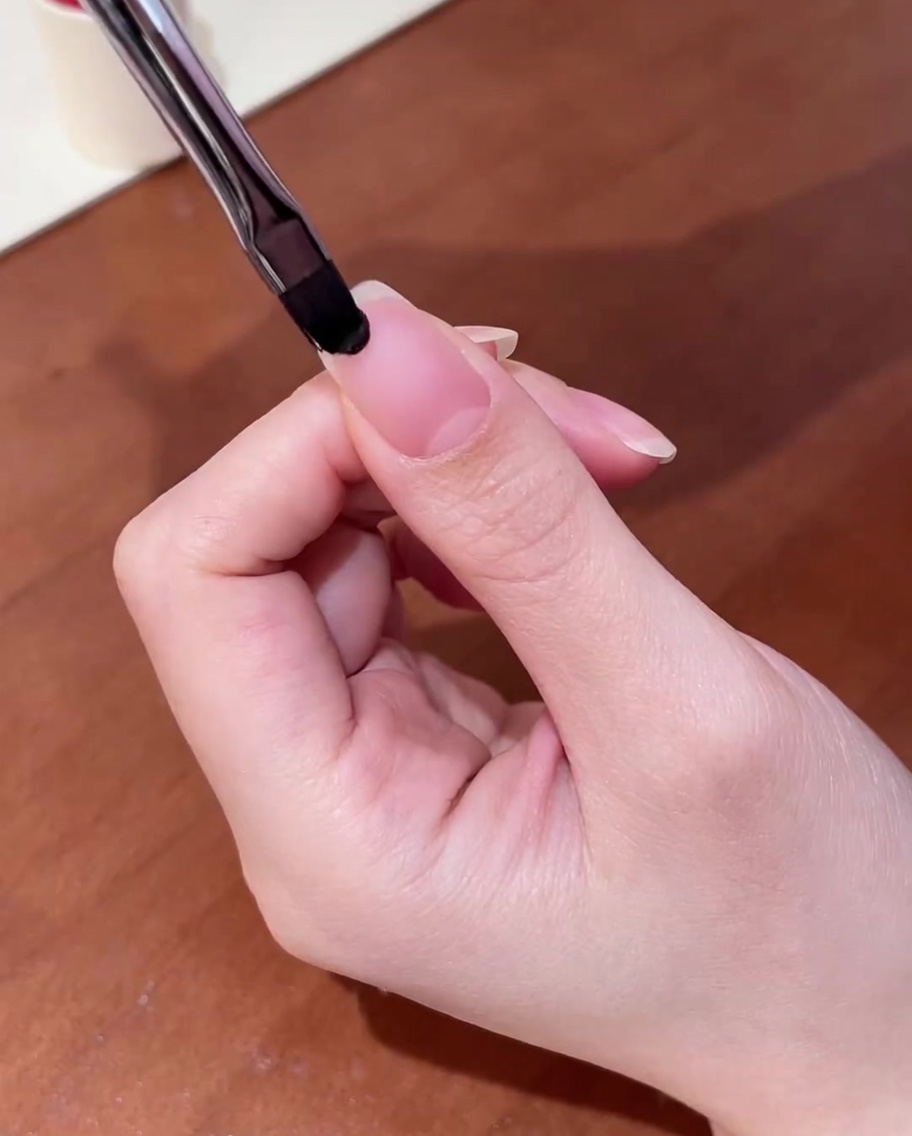 Primer plano de un pincel aplicando una capa base de gel transparente sobre una uña natural limpia.