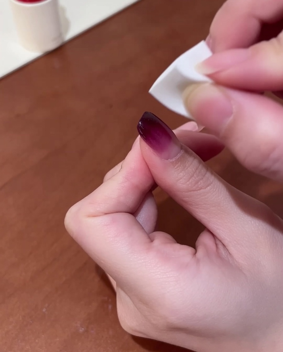 Primer plano de una mano sosteniendo una pequeña esponja de maquillaje, aplicando suavemente esmalte púrpura sobre una uña para crear un efecto degradado.