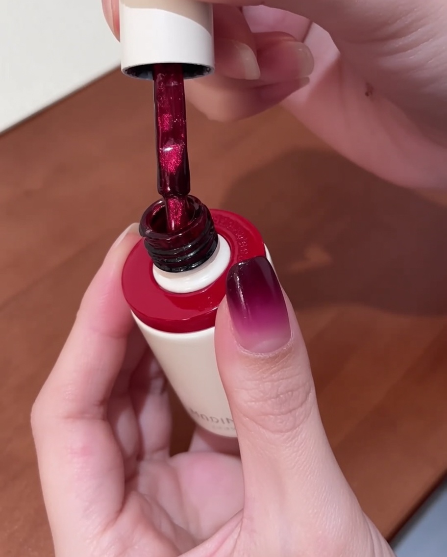 Un pincel aplicando esmalte en gel cat-eye rojo vibrante sobre una uña que ya tiene una base degradada.