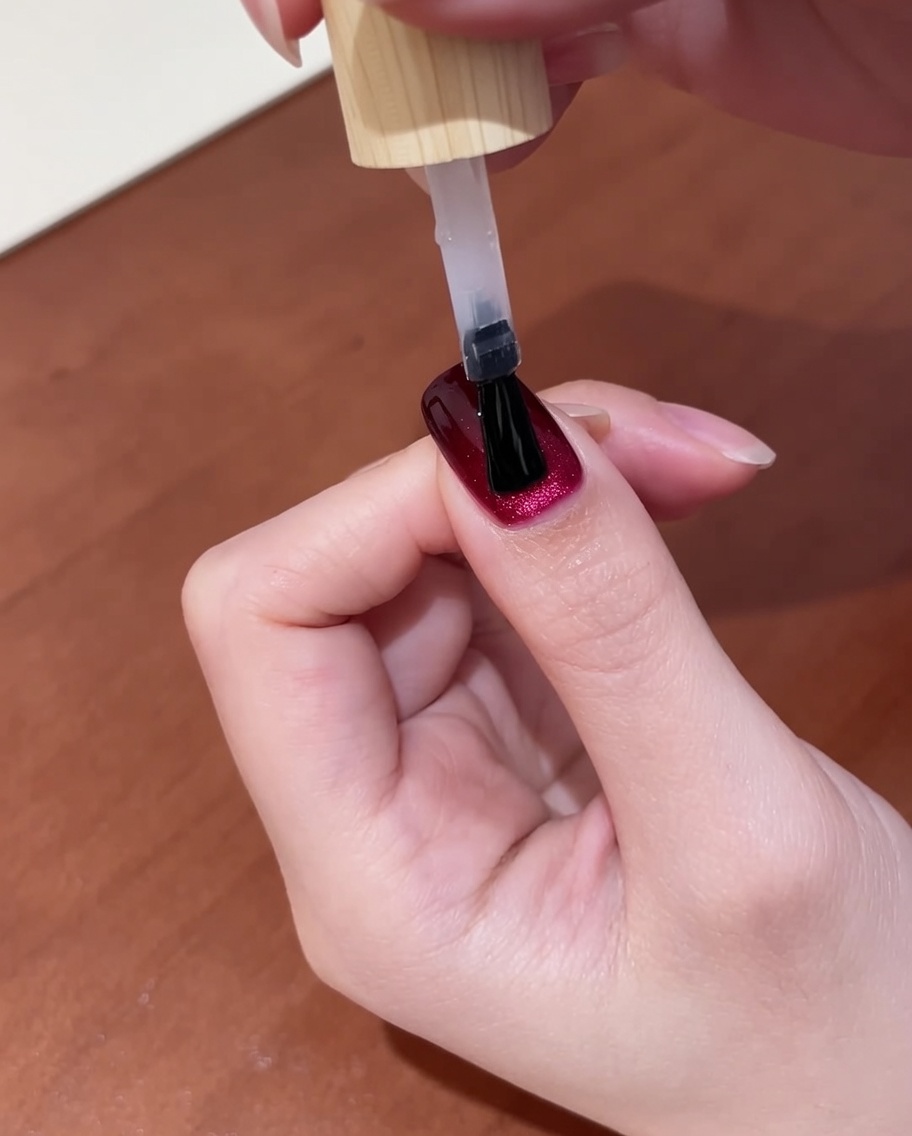 Vista cercana de una mano con top coat recién aplicado sobre uñas cat-eye rojo oscuro, colocada dentro de una lámpara UV para el paso final de curado.