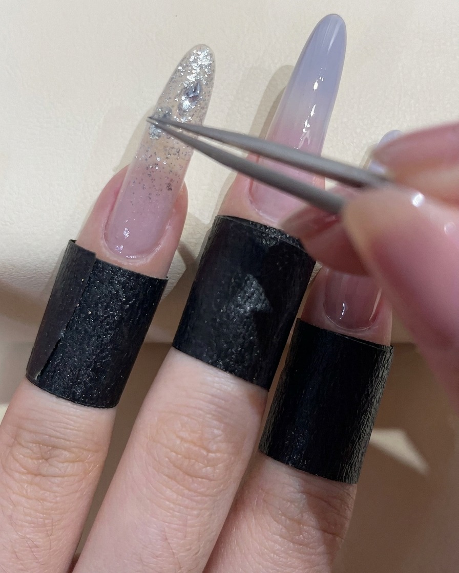 Unas pinzas colocando una pedrería transparente brillante sobre una uña cubierta con gel adhesivo húmedo.