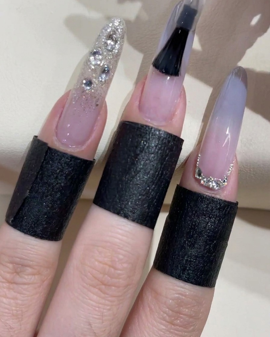 Un primer plano de un pincel de esmalte de uñas negro aplicando activamente un top coat transparente a una uña ombré azul claro, con los dedos adyacentes terminados mostrando detalles de purpurina plateada y pedrería.