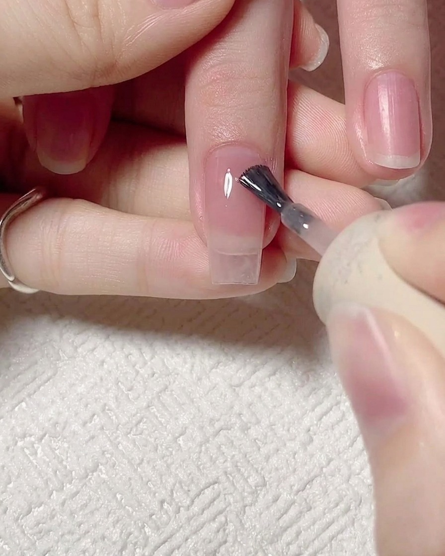 Primer plano de un pincel de uñas aplicando un gel jelly rosa translúcido sobre una uña natural limpia y preparada.