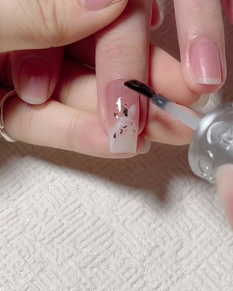 Un pincel aplicando base gel transparente sobre una uña con lámina de oro y diseños de ondas.