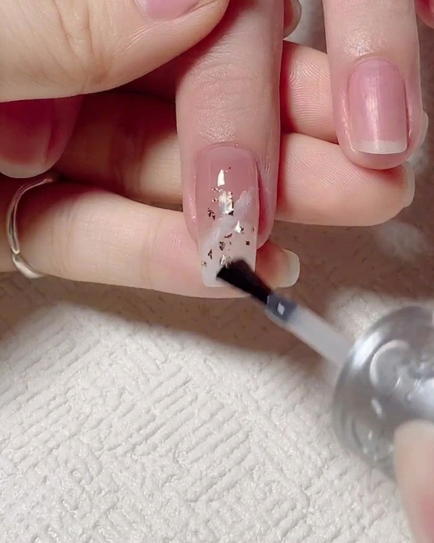Un pincel aplicando un top coat brillante final sobre la uña decorada, seguida de la mano bajo una lámpara UV.