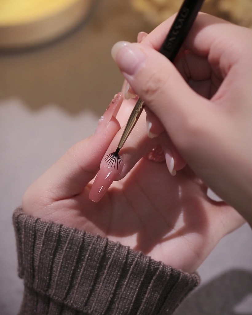 Primer plano de un pincel delineador siendo usado para delinear cuidadosamente el área de la cutícula con esmalte de gel rosa.