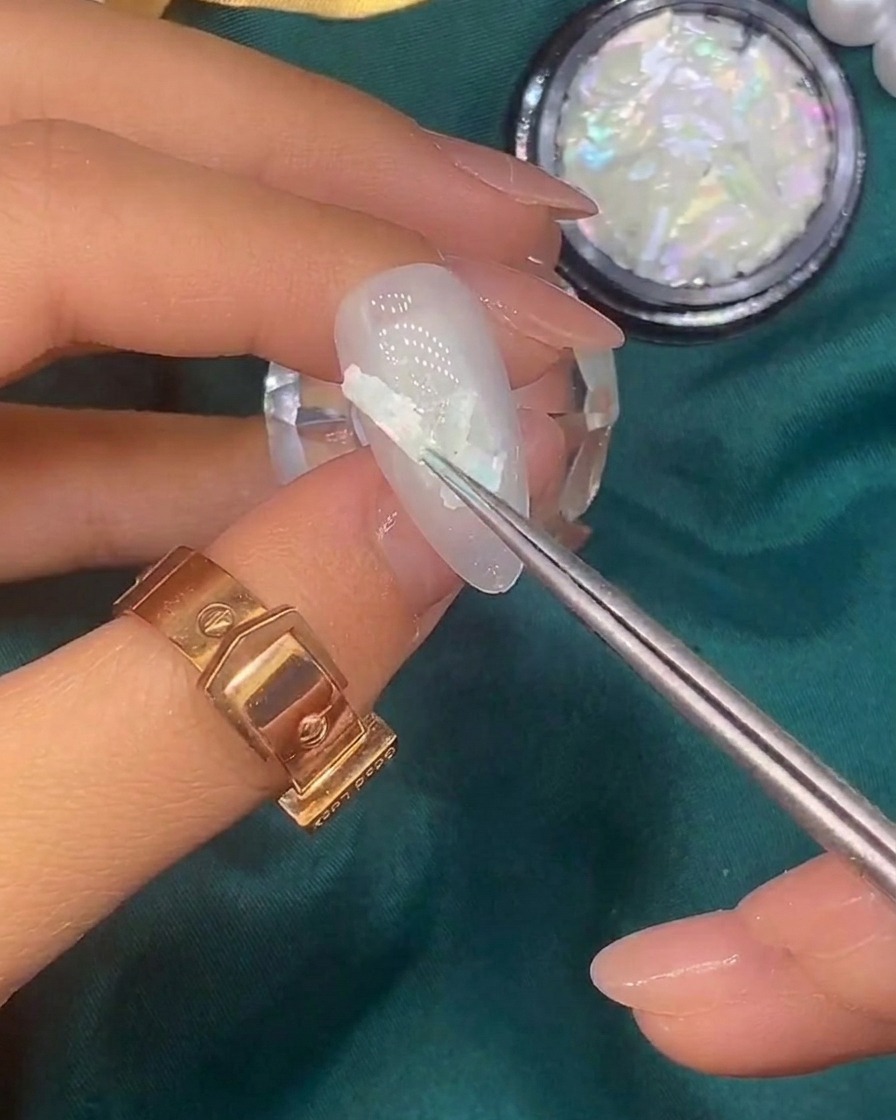 Un pincel aplicando pequeños copos de lámina dorada sobre la superficie de una uña decorada con conchas.