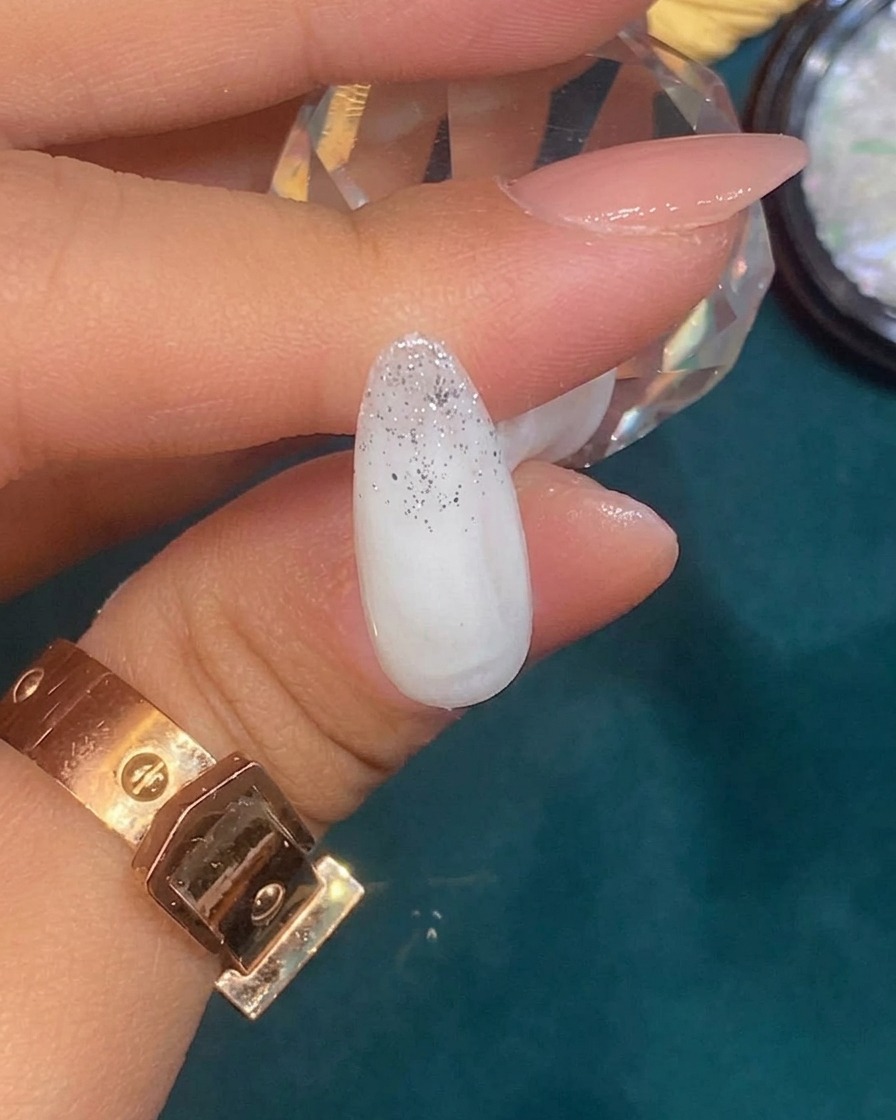 Pinzas colocando un pequeño trozo irregular de lámina plateada sobre una uña con degradado de glitter.