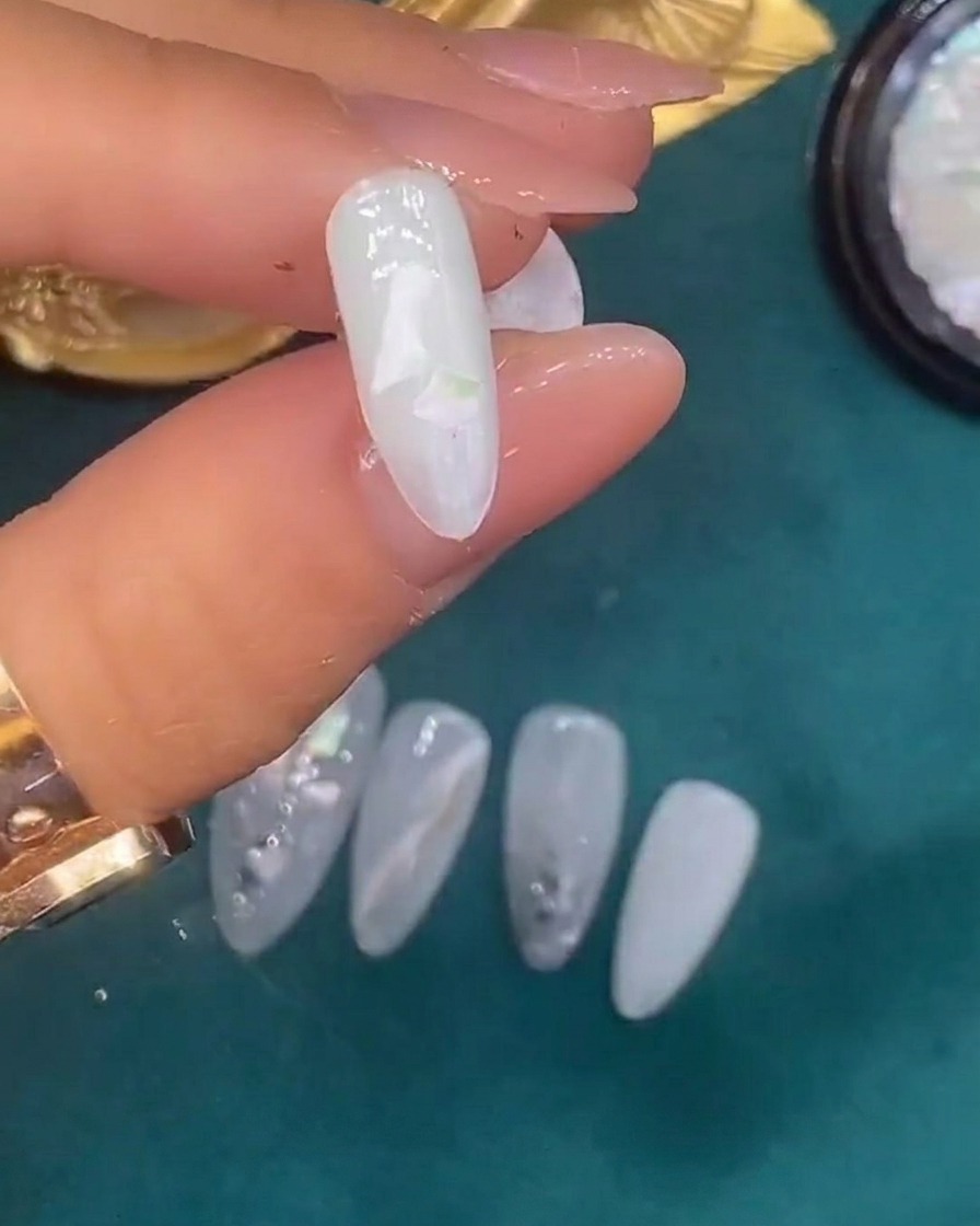 Pinzas colocando piezas decorativas de concha sobre una uña recubierta con gel blanco translúcido húmedo.