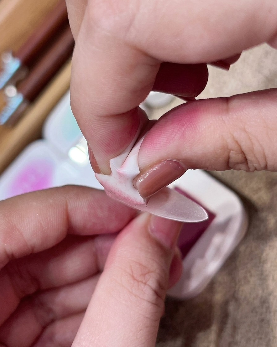 Una mano usando una pequeña esponja de maquillaje para aplicar pigmento rosa a toquecitos en el centro de una uña nude mate.