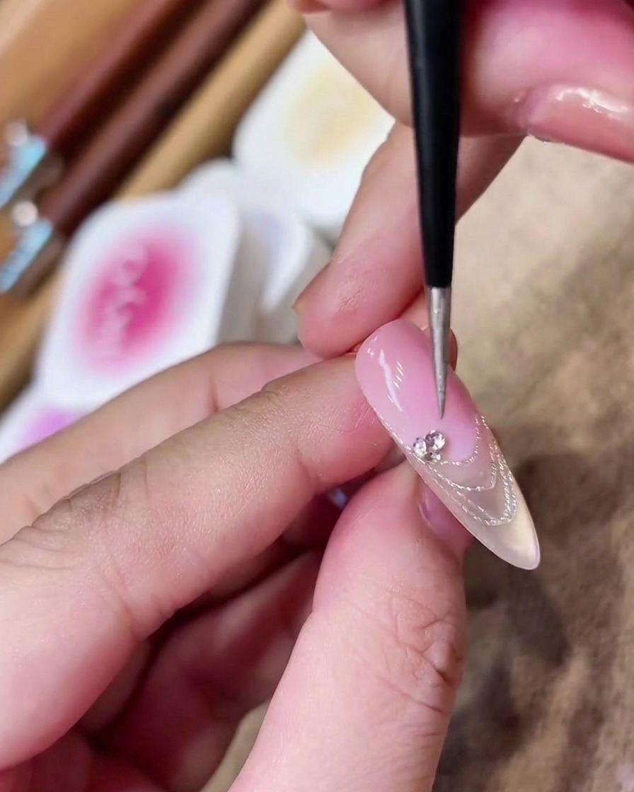 Un pincel de detalle colocando un pequeño grupo de cristales brillantes sobre la línea de la sonrisa de una manicura francesa.