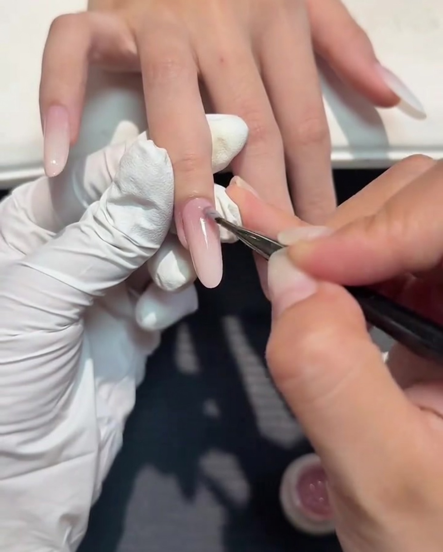 Un pequeño pincel de arte de uñas aplicando una capa delgada de gel nude sobre un degradado blanco lechoso.
