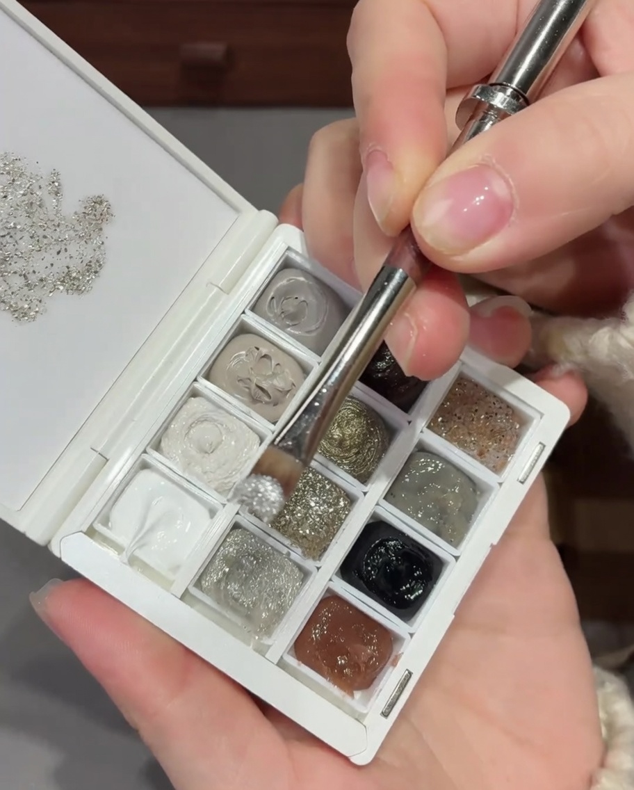 Un pincel de uñas aplicando una mezcla de purpurina y lentejuelas plateadas en la base de una uña natural.