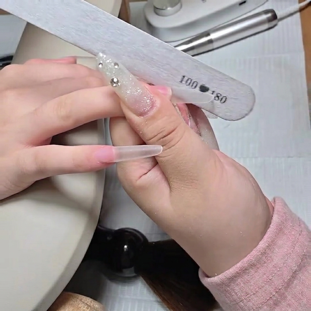 Primer plano de una lima manual dando forma al borde libre de un tip stiletto transparente largo.