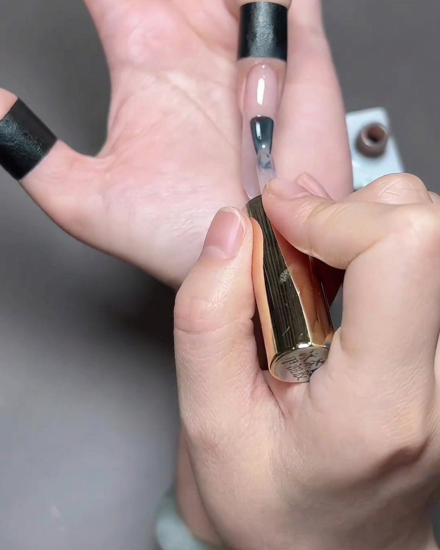 Un pincel aplicando un esmalte en gel translúcido de tono marrón claro en una uña natural larga.