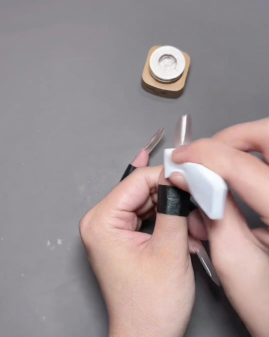 Primer plano de una mano presionando una plantilla en forma de corazón hueco sobre la uña del pulgar con un aplicador de esponja blanco, mientras la piel circundante está protegida con cinta protectora negracite: 1.