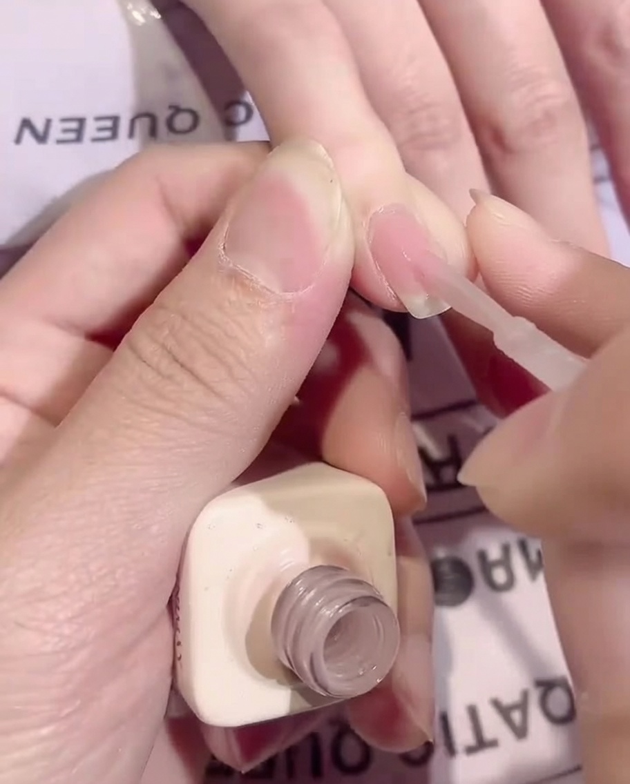 Un pincel aplicando una capa delgada y uniforme de gel base nude transparente sobre una uña.