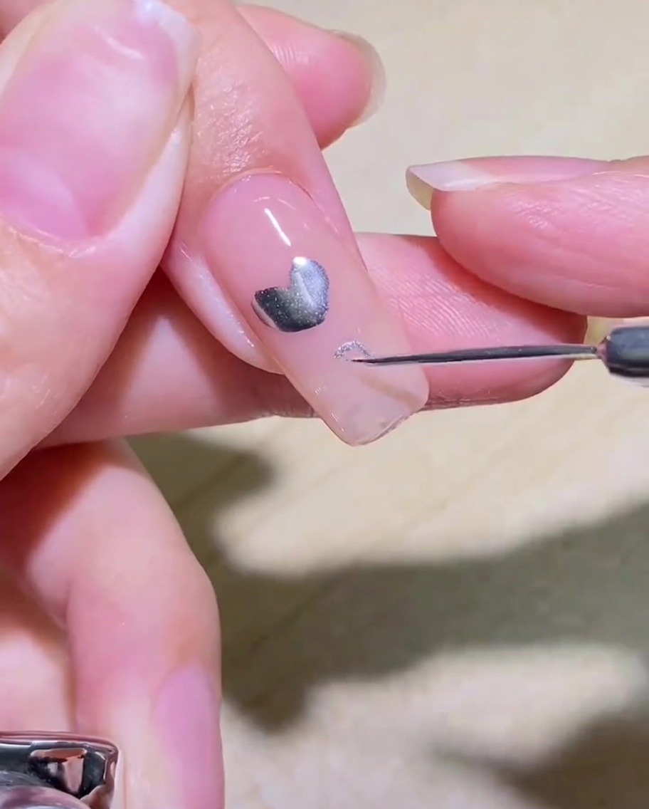 Primer plano de un pincel delineador dibujando el contorno de un corazón plateado sobre una uña transparente.