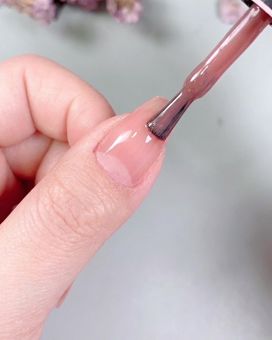Un pequeño pincel plano para gel alisando suavemente el color nude sobre la superficie de la uña.