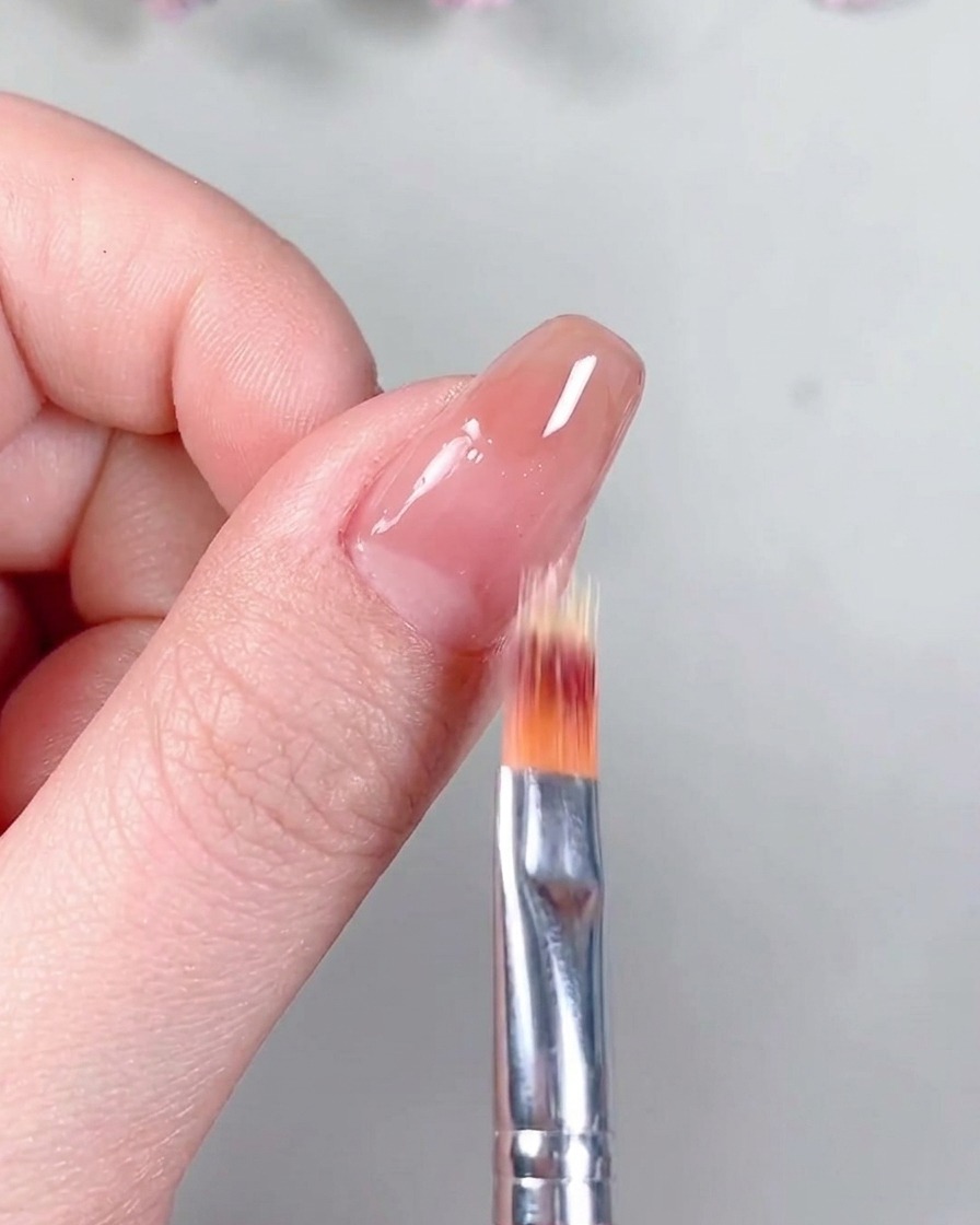 Un pincel aplicando un top coat transparente de acabado mate sobre la base de gel nude ya curada.