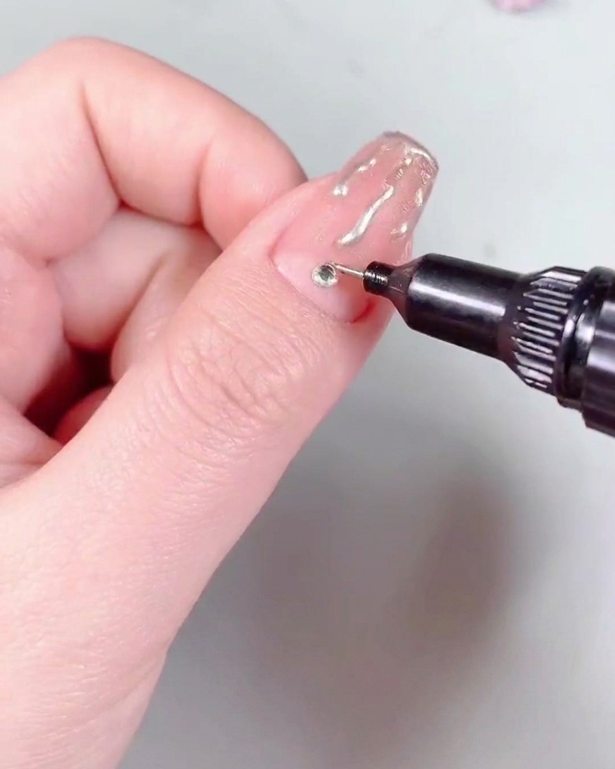 Un pincel de nail art aplicando una capa de top coat de alto brillo sobre una uña terminada con el diseño de llamas y un cristal.