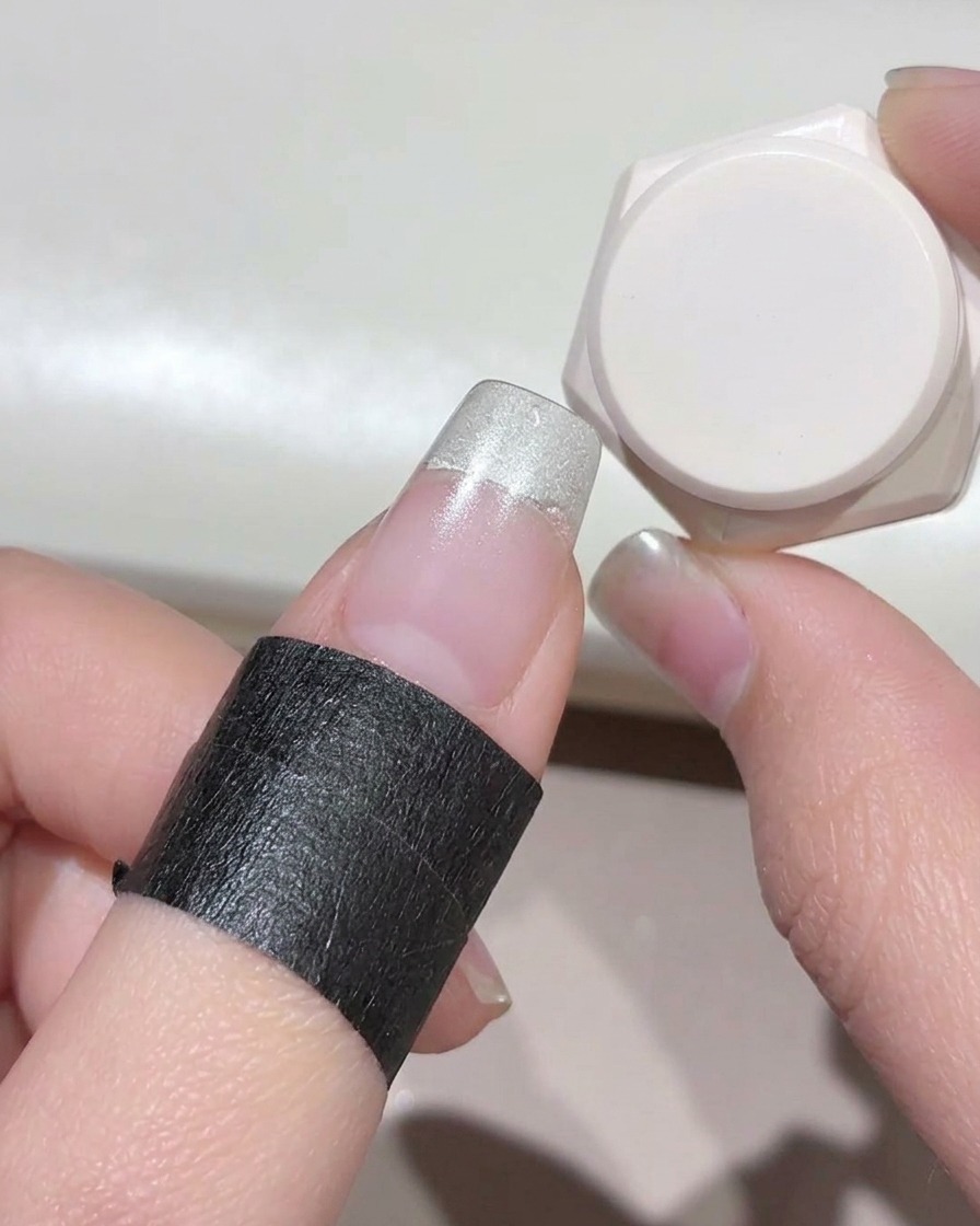 Primer plano de un dedo con cinta protectora negra alrededor de la piel, mostrando gel blanco lechoso aplicado en la punta de la uña.