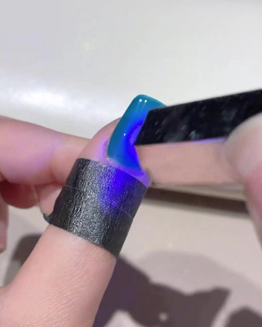 Una mini lámpara UV de mano proyectando luz azul sobre una uña con gel ojo de gato azul profundo para curar el diseño.