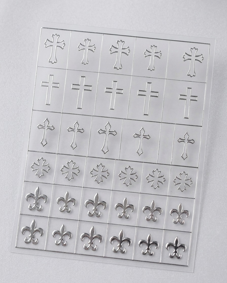 Primer plano que muestra unas pinzas de punta fina levantando una delicada pegatina de plantilla en forma de cruz de color plateado de una hoja adhesiva transparente.
