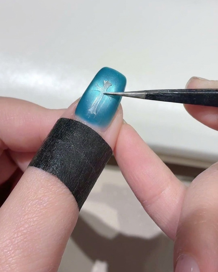 Primer plano de unas pinzas presionando una pegatina de plantilla en forma de cruz transparente sobre el centro de una uña azul brillante con efecto ojo de gato.