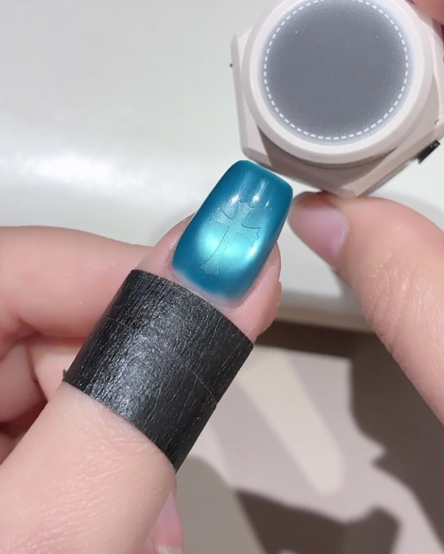 Una pequeña herramienta de esponja redonda flotando sobre una uña azul que tiene una pegatina en forma de cruz, preparándose para aplicar esmalte en gel negro.