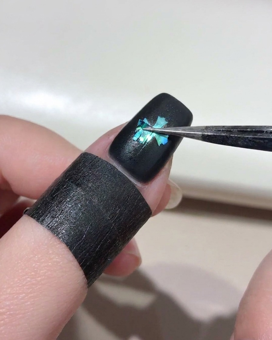 Unas pinzas levantando cuidadosamente una plantilla de cruz de una uña pintada de negro, revelando una forma de cruz azul brillante con efecto ojo de gato debajo.