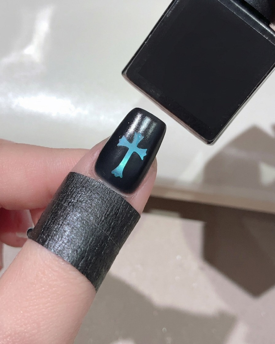 Un pincel aplicando un top coat transparente y brillante sobre una uña negra con un brillante diseño de cruz azul en el centro.