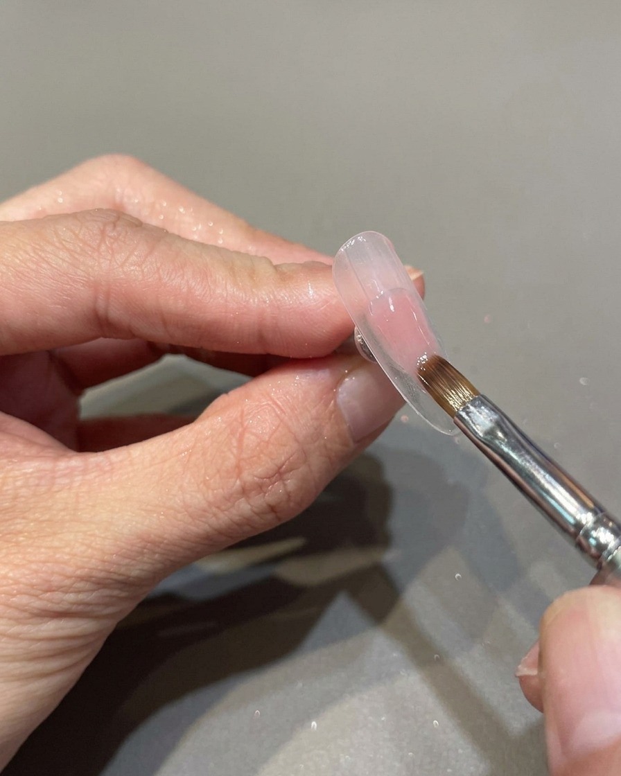Pincel profesional aplicando una capa de esmalte de gel rosa traslúcido sobre la superficie de una uña preparada.
