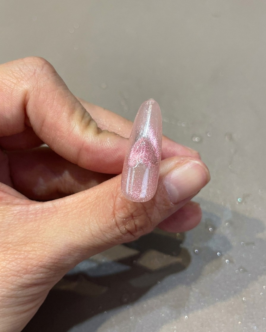 Primer plano de una uña rosa con diseño de corazón cat-eye, mostrando el uso de un imán para manipular el efecto brillante.