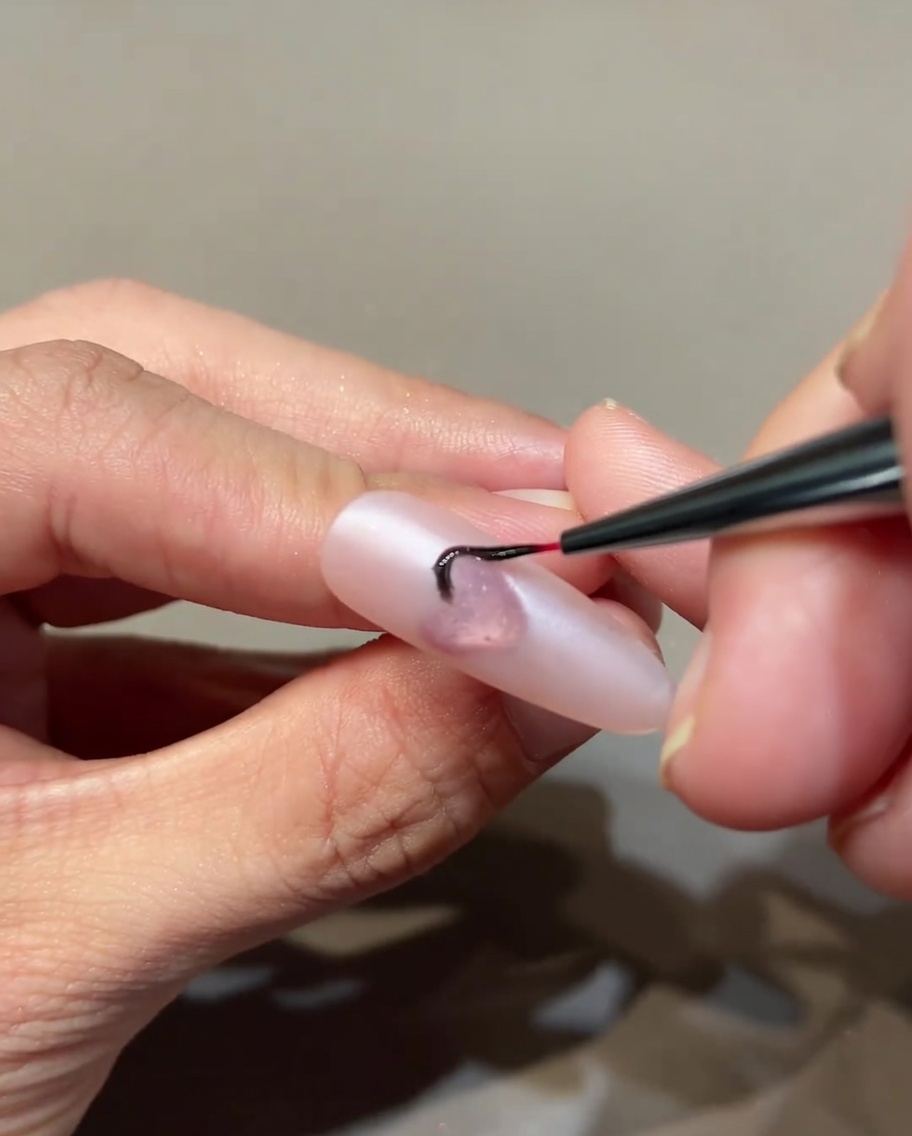 Un pincel fino de nail art dibujando detalles finos de llamas negras sobre una base rosa alrededor de un corazón central.