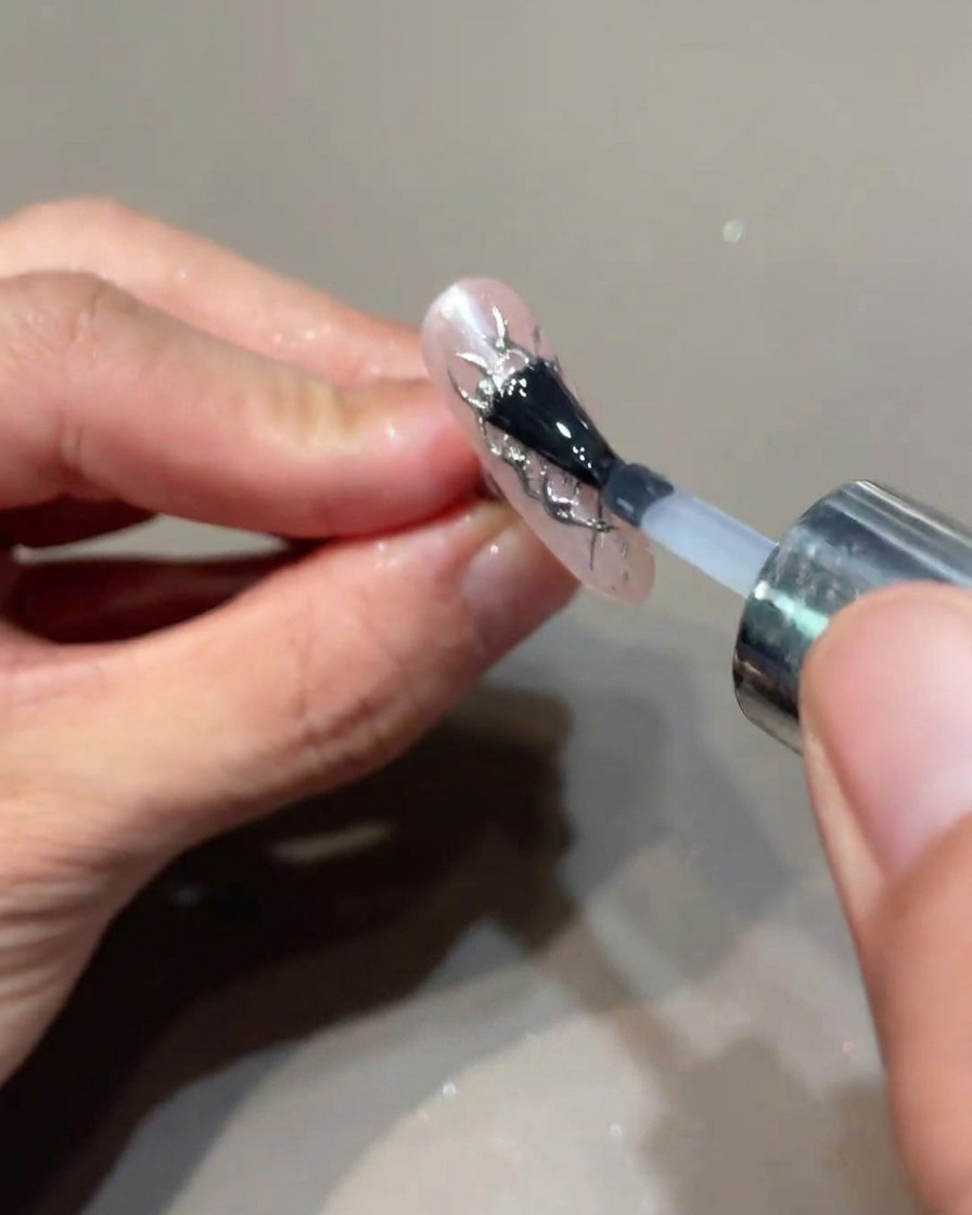 Un pincel aplicando una capa final de top coat transparente sobre una uña decorada con acentos de llamas plateadas.