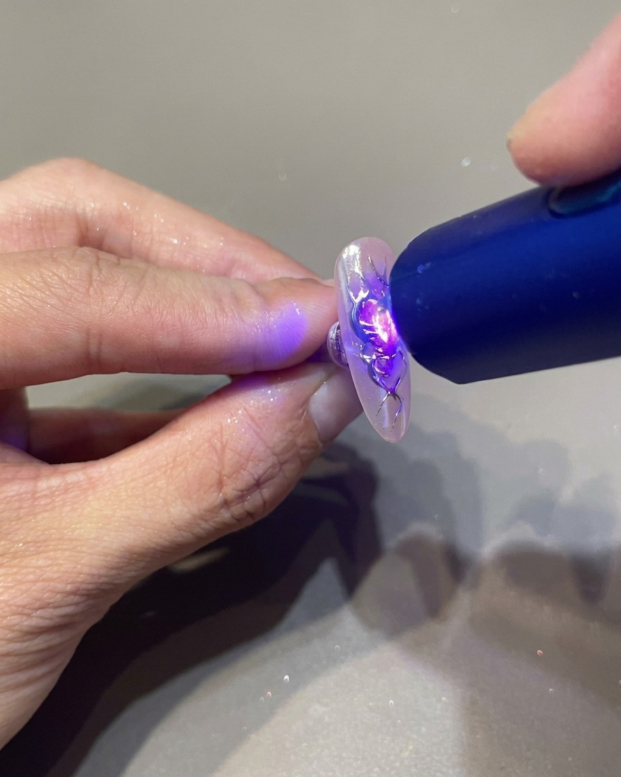 Una lámpara UV portátil sostenida sobre una uña decorada para fijar los detalles de gel 3D transparente en un diseño de corazón con llamas.